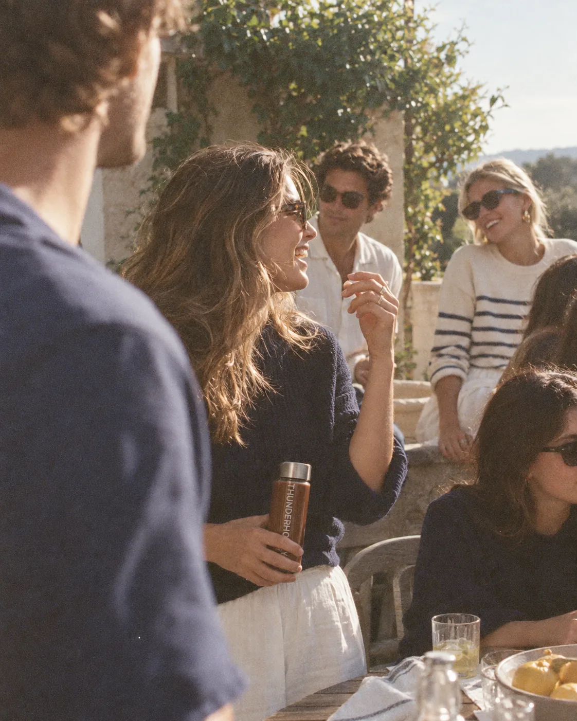 Friends laughing on a sunny terrace by the sea, one of them holding a bottle of the drink