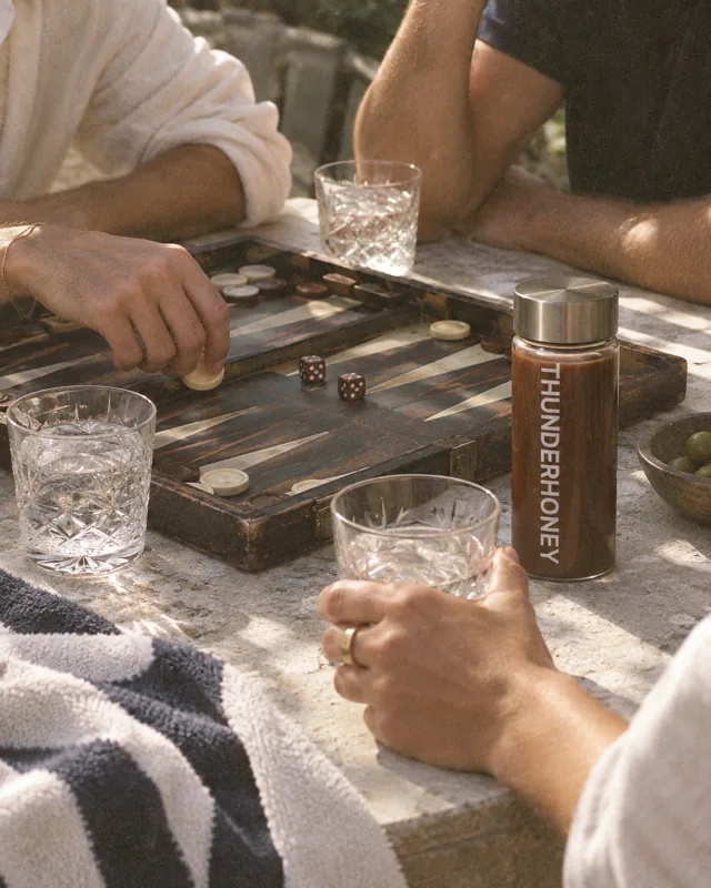 Hands over a backgammon board mid game, glasses of water and a Thunder Honey bottle beside it