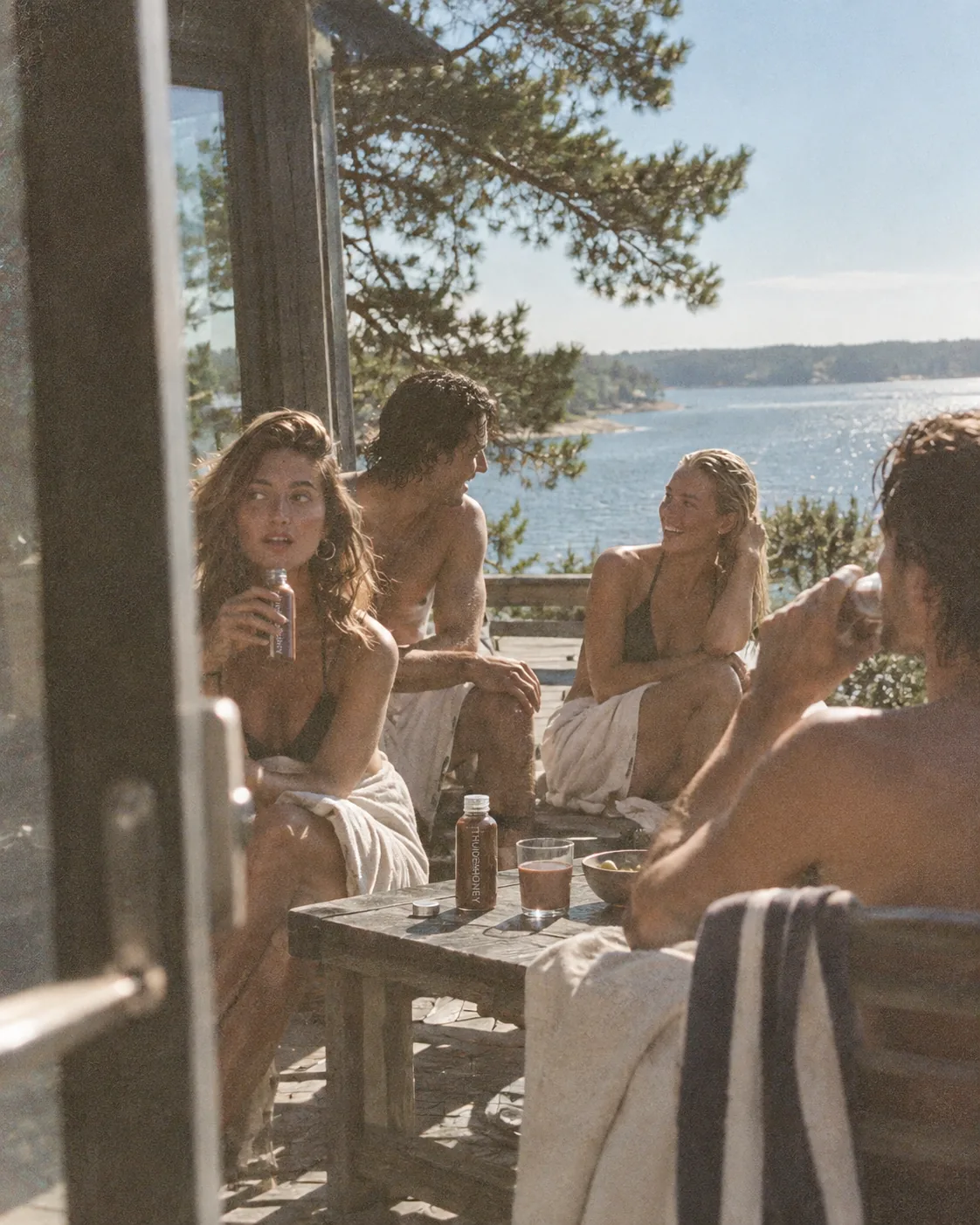 Friends in towels around a table by the water after a swim, cups of the drink between them