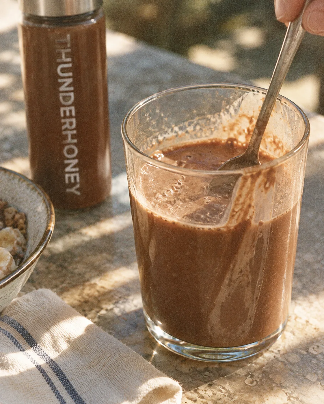 A glass of Thunder Honey cacao being stirred with a spoon, the bottle beside it in the sun