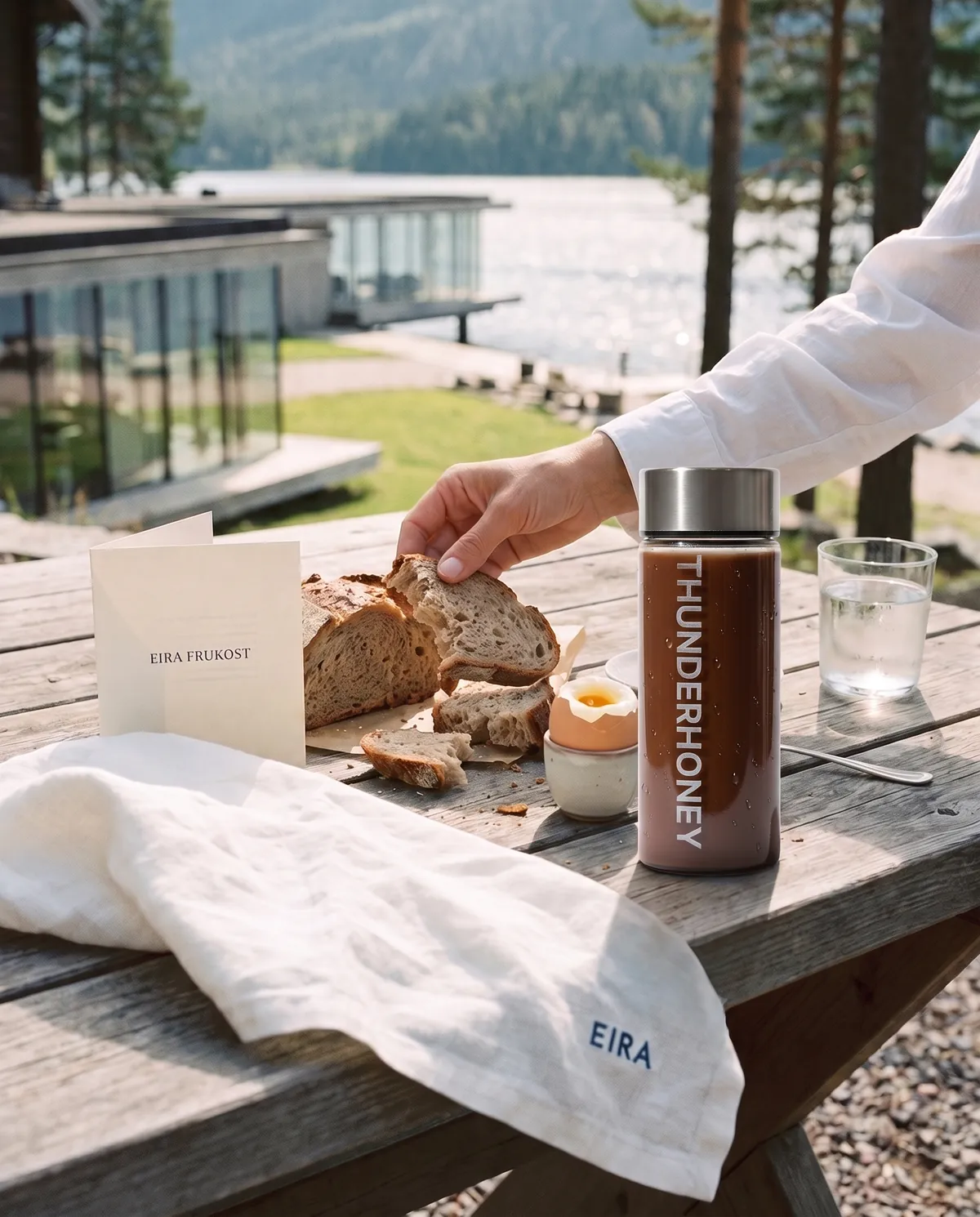 A Thunder Honey glass bottle filled with the cacao drink on an outdoor breakfast table by a lake, next to fresh bread and a soft-boiled egg
